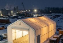 White fabric storage shelter lit from inside, with an open roll-up door, on a snowy industrial yard at night beside crates and equipment