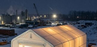 White fabric storage shelter lit from inside, with an open roll-up door, on a snowy industrial yard at night beside crates and equipment