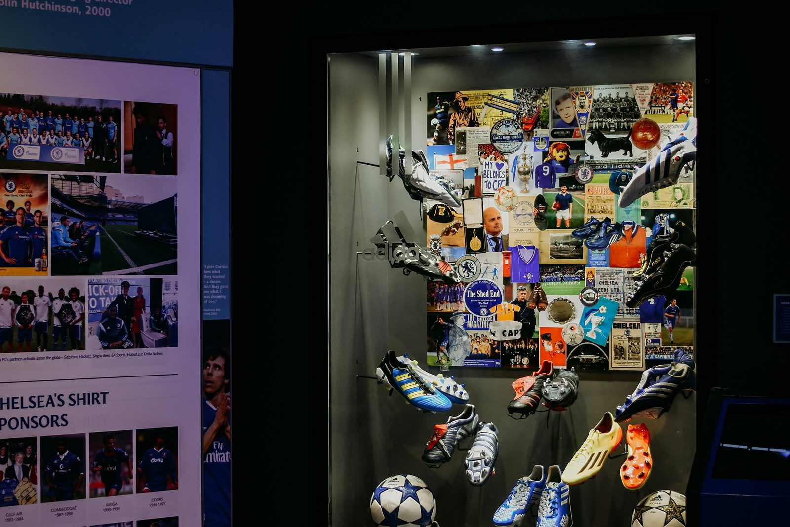Display case of adidas football boots and balls in front of a collage of Chelsea FC memorabilia, beside a panel on Chelsea’s shirt sponsors