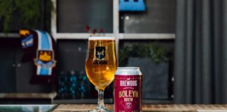 A can of BrewDog Boleyn Brew oat lager beside a full BrewDog glass on a wooden table, with a West Ham United scarf in the background