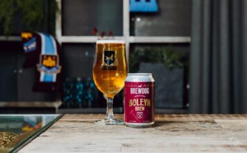 A can of BrewDog Boleyn Brew oat lager beside a full BrewDog glass on a wooden table, with a West Ham United scarf in the background