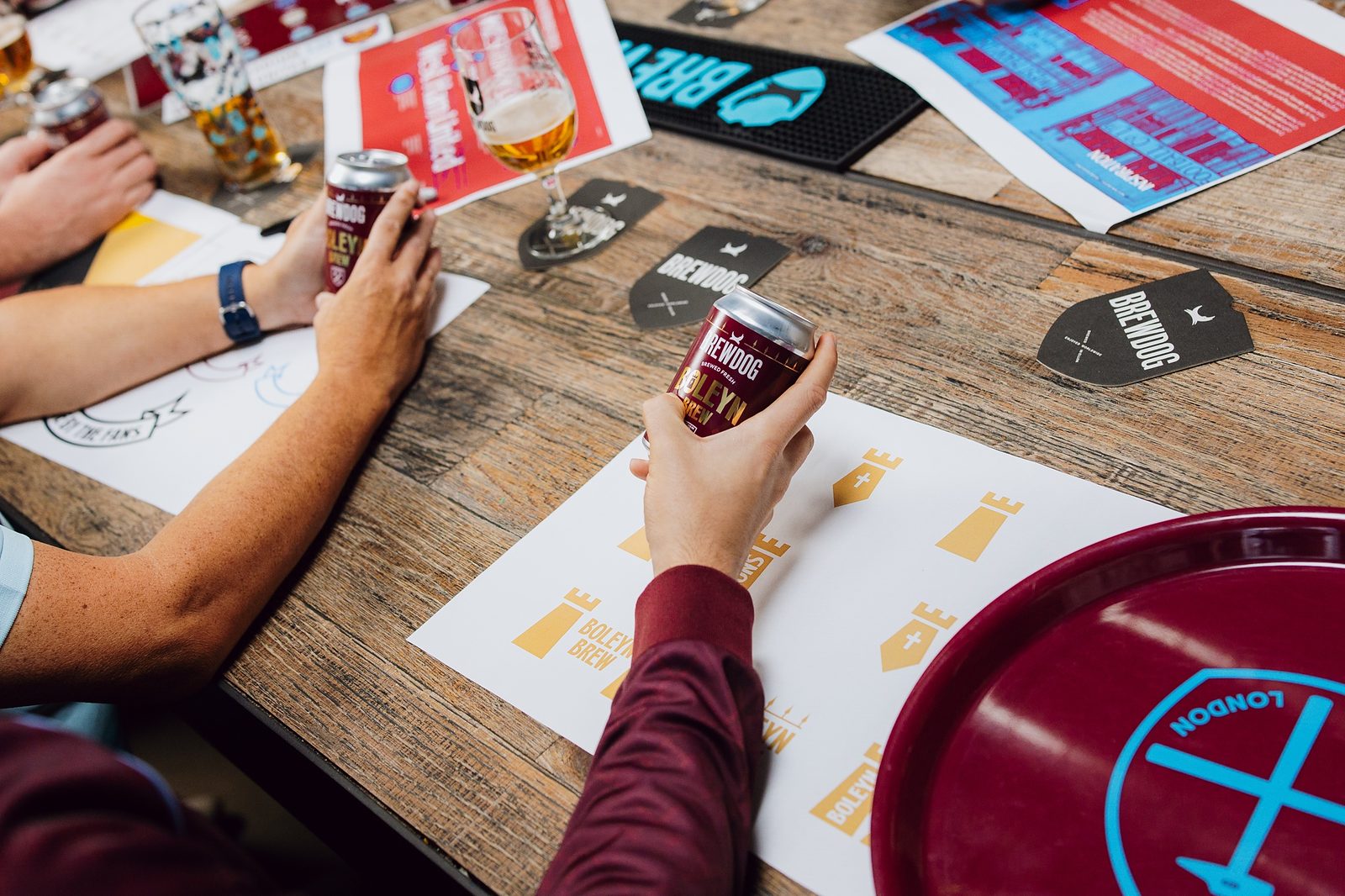 Hands holding cans of Boleyn Brew over printed Boleyn Brew logo designs and BrewDog beer mats