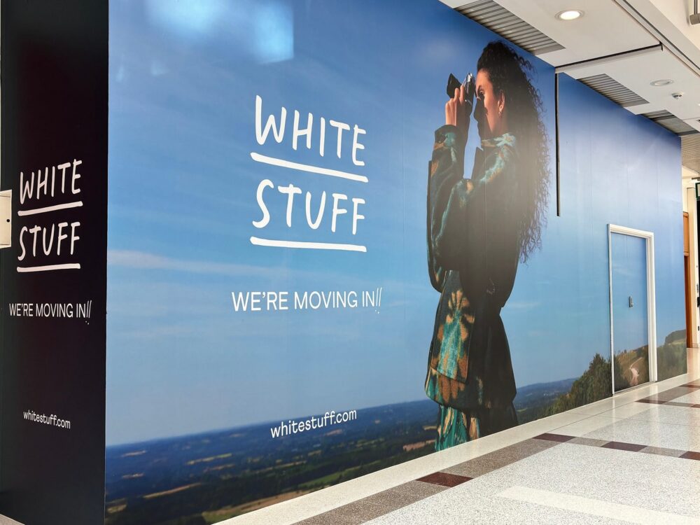 White Stuff store hoarding in a shopping centre mall reading We’re moving in, with a woman holding a camera against a countryside backdrop