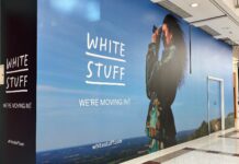 White Stuff store hoarding in a shopping centre mall reading We’re moving in, with a woman holding a camera against a countryside backdrop