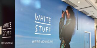 White Stuff store hoarding in a shopping centre mall reading We’re moving in, with a woman holding a camera against a countryside backdrop