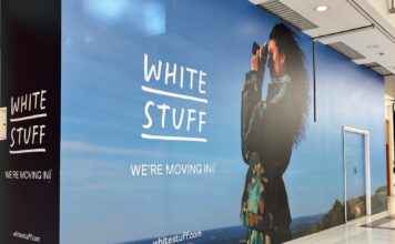 White Stuff store hoarding in a shopping centre mall reading We’re moving in, with a woman holding a camera against a countryside backdrop